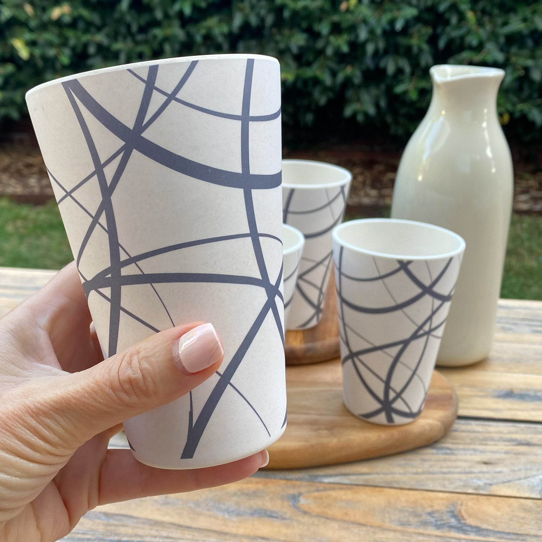 Bamboo cup with abstract design held by a hand, with more cups and a pitcher on a wooden surface outdoors.