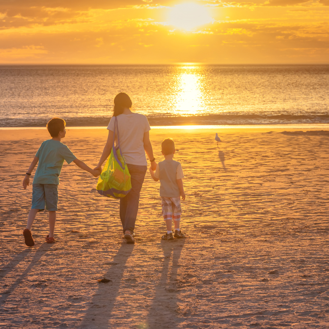 a mother holding her 2 sons hands on the beach walking towards the water with the sunsetting in the background.