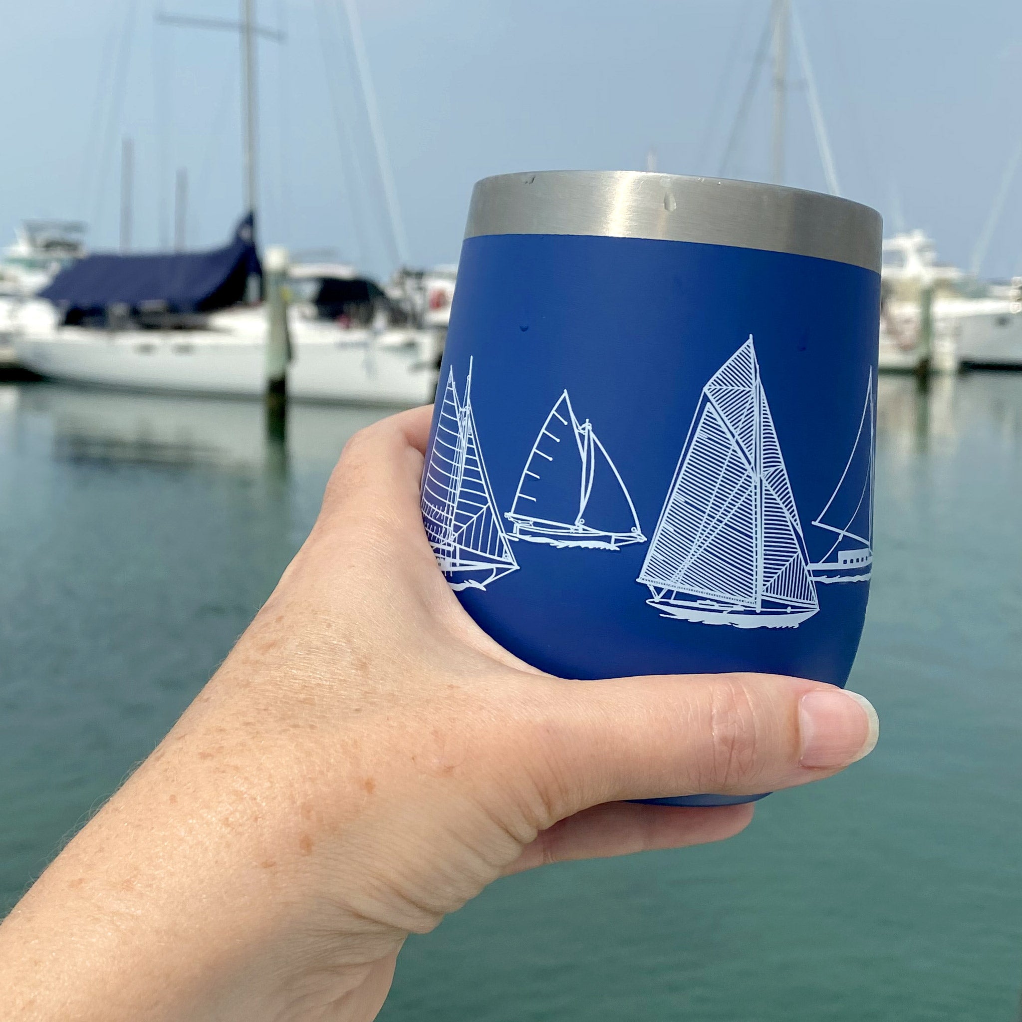 A lady's hand holding a blue Van Go Collections NautiGo Insulated Wine Cup with boats in the background