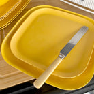 bamboo plate 22cm on 26cm in sunshine yellow, on table with knife, angled